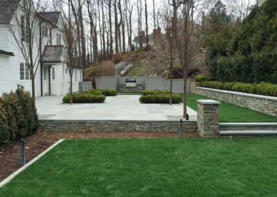 Modern residential patio and surround renovation features stone patio, stacked stone retaining wall and steps, and plantings.
