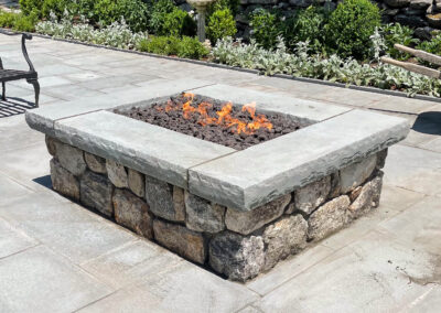 Contemporary residential square, stacked natural stone fire pit with thick stone coping installed on a polished stone patio.